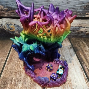 May include: A rainbow-coloured 3D printed dragon head dice tower with a purple base and a purple, blue, and green dragon head. The dragon head is open and there are four dice inside the tower.