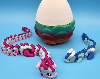 Può includere: Tre giocattoli a forma di drago colorati, uno rosa e blu, uno blu e verde e uno in posizione arrotolata. I draghi sono tutti in plastica e hanno un corpo segmentato. I draghi sono seduti su una superficie blu davanti a un uovo bianco con una base verde e rossa.