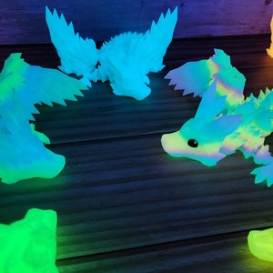 May include: A group of seven glow-in-the-dark dragon figurines in various shades of blue, green, and orange. The dragons are posed in different positions, with some flying and others resting. The figurines are made of a translucent material that allows the light to shine through.