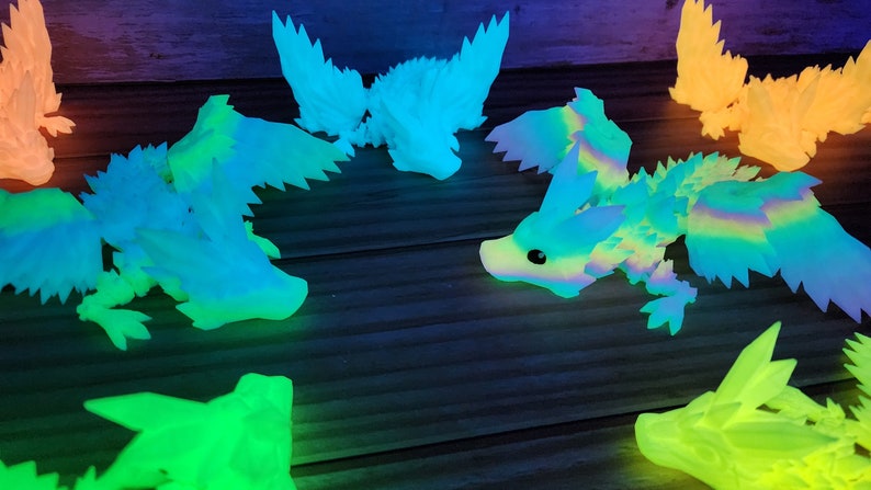 May include: A group of seven glow-in-the-dark dragon figurines in various shades of blue, green, and orange. The dragons are posed in different positions, with some flying and others resting. The figurines are made of a translucent material that allows the light to shine through.