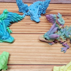 May include: A collection of six 3D printed dragon figurines. The dragons are all different colors, including blue, green, pink, and orange. They have spiky wings and tails and are posed in a variety of positions.