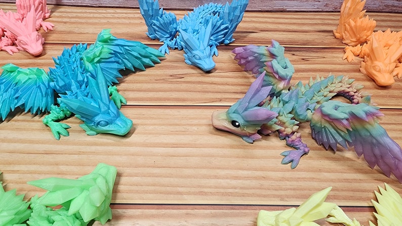 May include: A collection of six 3D printed dragon figurines. The dragons are all different colors, including blue, green, pink, and orange. They have spiky wings and tails and are posed in a variety of positions.