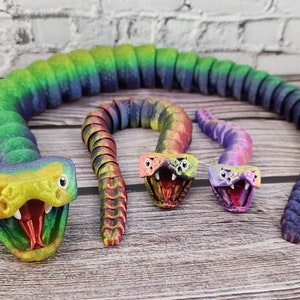May include: Three articulated snake toys in a rainbow gradient. The largest is fully extended, the others coiled. The snakes have detailed heads with open mouths and visible fangs. The toys are made of plastic.