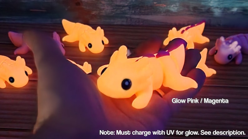 May include: Five glow-in-the-dark axolotl figurines in various poses. The axolotls are made of a soft, rubbery material and are painted in a bright pink colour. The text "Glow Pink / Magenta" and "Note: Must charge with UV for glow. See description." is visible in the image.