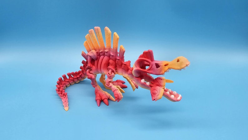 Pu&ograve; includere: Un modello di scheletro di dinosauro stampato in 3D con un gradiente di colore rosso brillante e arancione. Il dinosauro &egrave; in posizione di corsa con la bocca aperta e la coda arrotolata.