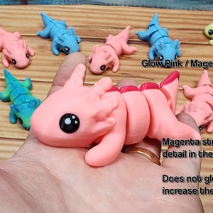 May include: A collection of small, segmented, colourful, plastic toys resembling axolotls. The toys are in various colours, including pink, blue, green, and yellow. The pink axolotl has a magenta stripe down its back. The text "Glow Pink / Magenta" and "Magenta stripe adds beautiful detail in the light." is visible in the image. The text "Does not glow in the dark to increase the effect" is also visible.