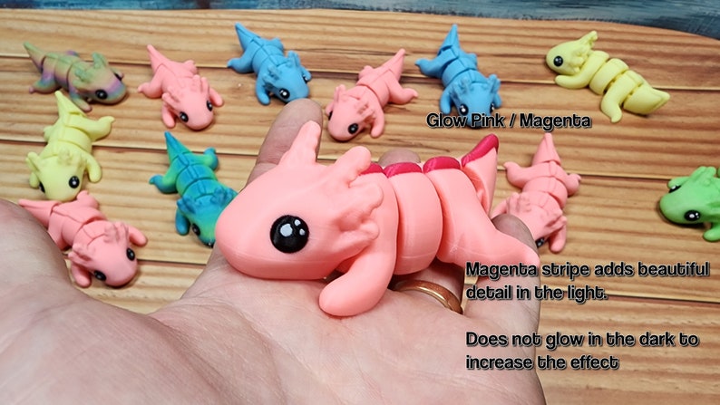 May include: A collection of small, segmented, colourful, plastic toys resembling axolotls. The toys are in various colours, including pink, blue, green, and yellow. The pink axolotl has a magenta stripe down its back. The text "Glow Pink / Magenta" and "Magenta stripe adds beautiful detail in the light." is visible in the image. The text "Does not glow in the dark to increase the effect" is also visible.