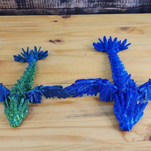 Może przedstawiać: Dwa smoki origami, jeden niebieski, a drugi zielony, z rozłożonymi skrzydłami. Smoki są wykonane z papieru i mają szczeg&oacute;łową, łuskowatą fakturę.