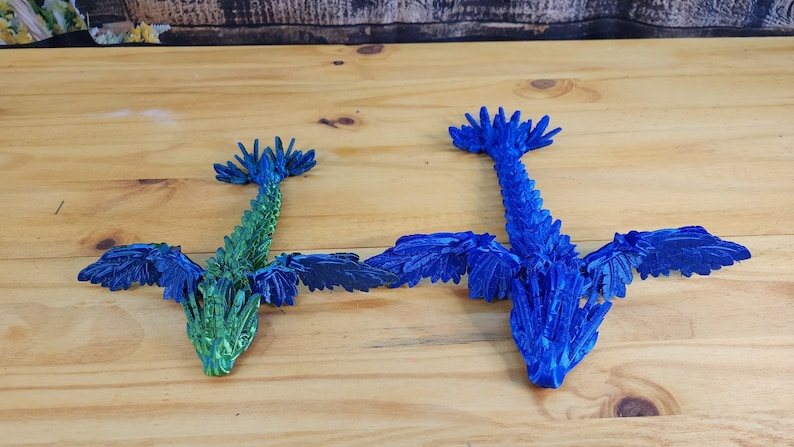 Może przedstawiać: Dwa smoki origami, jeden niebieski, a drugi zielony, z rozłożonymi skrzydłami. Smoki są wykonane z papieru i mają szczeg&oacute;łową, łuskowatą fakturę.
