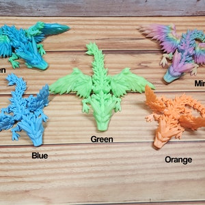 May include: A set of 7 colorful dragon figurines, each with a different color: blue/green, blue, green, orange, pink, yellow, and rainbow. The dragons are made of a textured material and have wings and tails.