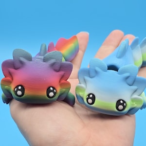 May include: Two small axolotl figurines, one with a rainbow gradient and the other with a blue and green gradient. Both have large eyes and a cute expression.