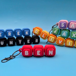 May include: A collection of colorful silicone cube bead keychains featuring the names Derek, Hunter, Sam, Jax, Olivia, and Ben. Each keychain consists of square beads with white letters, some including star or cat icons, attached to metal clasps.