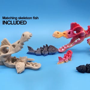 Pu&ograve; includere: Un set di giocattoli scheletrici articolati, tra cui un dinosauro rosa e arancione, un pesce grigio e un pesce nero. Il testo "Matching skeleton fish INCLUDED" &egrave; visibile nell'immagine.