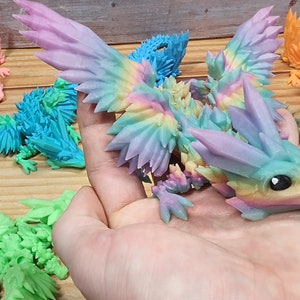 May include: A rainbow-colored, crystal-like dragon figurine with wings outstretched. The dragon is made of a 3D printed material and has a detailed, textured surface.