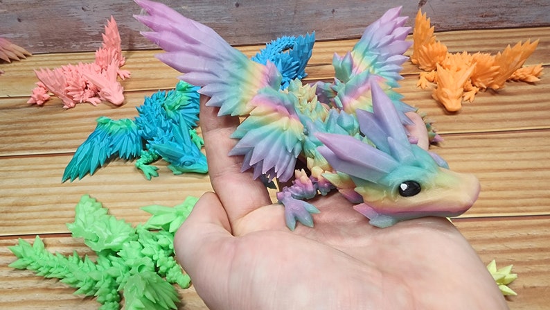 May include: A rainbow-colored, crystal-like dragon figurine with wings outstretched. The dragon is made of a 3D printed material and has a detailed, textured surface.