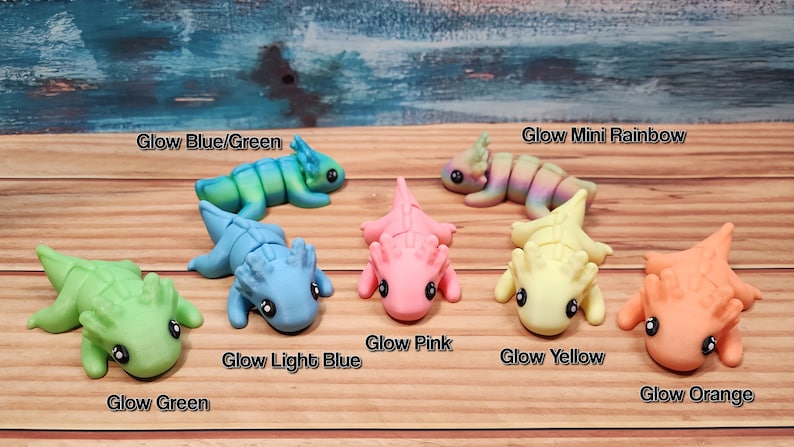 May include: A set of seven glow-in-the-dark miniature axolotl figurines in various colours, including green, light blue, pink, yellow, orange, and a rainbow colour. The figurines are arranged on a wooden surface.