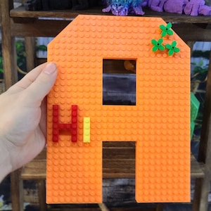 May include: An orange building block construction with a window and a roof. The word "Hi" is spelled out in red and yellow blocks. Green flower-shaped blocks are attached to the roof. The building block is held by a hand.