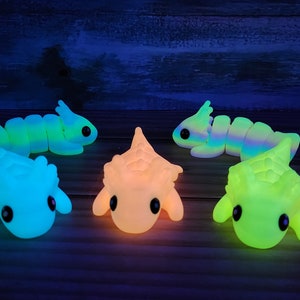 May include: A group of seven glow-in-the-dark toys. The toys are shaped like various creatures, including a worm, a dinosaur, and a hedgehog. The toys are all different colours, including green, blue, yellow, and orange.