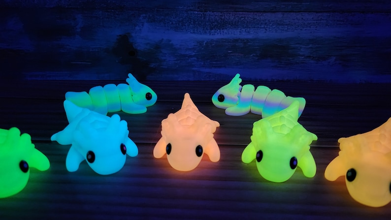 May include: A group of seven glow-in-the-dark toys. The toys are shaped like various creatures, including a worm, a dinosaur, and a hedgehog. The toys are all different colours, including green, blue, yellow, and orange.