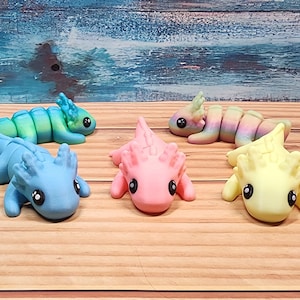 May include: Seven colourful axolotl figurines, each with a different colour scheme. The axolotls are posed in a variety of positions, some are lying down, some are sitting up, and some are standing. The figurines are made of a smooth, glossy material and have a realistic appearance.