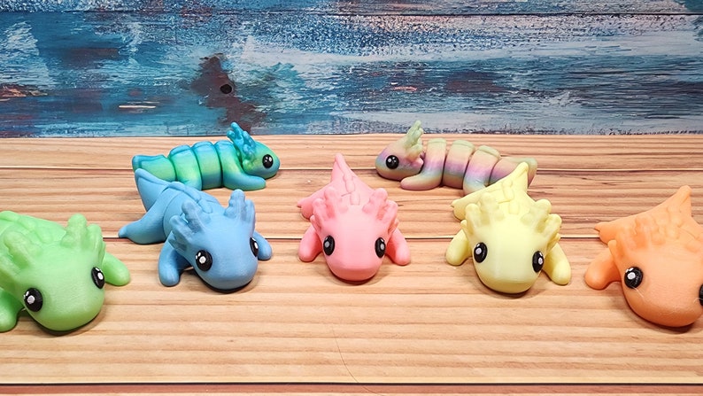 May include: Seven colourful axolotl figurines, each with a different colour scheme. The axolotls are posed in a variety of positions, some are lying down, some are sitting up, and some are standing. The figurines are made of a smooth, glossy material and have a realistic appearance.