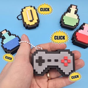 May include: A collection of pixel art keychains featuring a gray game controller, a yellow coin, and three potion bottles in light blue, light green, and red. Each item has a black border and a metal ball chain attached for hanging purposes.