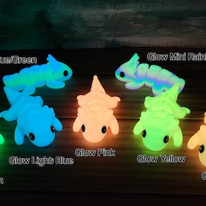 May include: Seven glow-in-the-dark dinosaur toys in various colours, including green, blue, pink, yellow, and orange. The dinosaurs are arranged in a semi-circle on a wooden surface.