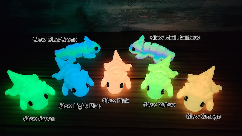 May include: Seven glow-in-the-dark dinosaur toys in various colours, including green, blue, pink, yellow, and orange. The dinosaurs are arranged in a semi-circle on a wooden surface.