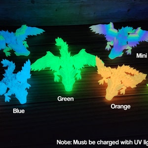 May include: A set of glow-in-the-dark dragon figurines in various colors, including pink, blue, green, orange, yellow, and a multi-colored rainbow dragon. The text "Note: Must be charged with UV light. See Description." is visible at the bottom of the image.