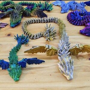 Może przedstawiać: Kolekcja tęczowych smok&oacute;w origami w r&oacute;żnych kolorach, w tym zielonym, niebieskim, fioletowym i złotym. Smoki są wykonane z papieru i mają skomplikowane detale.