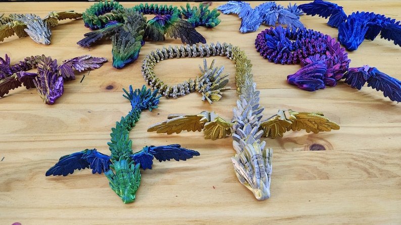 Może przedstawiać: Kolekcja tęczowych smok&oacute;w origami w r&oacute;żnych kolorach, w tym zielonym, niebieskim, fioletowym i złotym. Smoki są wykonane z papieru i mają skomplikowane detale.