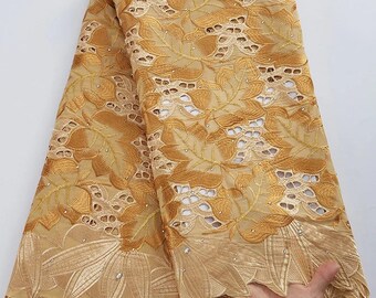 May include: Gold-colored lace fabric with a floral leaf design. The fabric has cut-out patterns and is embellished with small, sparkling accents. The edges feature a scalloped design. The fabric is folded over, showing the intricate details of the lace.