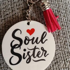 Op de afbeelding: Ronde witte sleutelhanger met de tekst "Soul Sister" in zwarte cursieve letters en een rood hart boven het woord "Soul". De sleutelhanger heeft een zilveren ketting en een roze kwast.