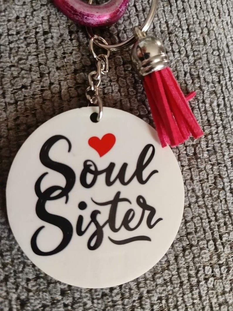 Op de afbeelding: Ronde witte sleutelhanger met de tekst "Soul Sister" in zwarte cursieve letters en een rood hart boven het woord "Soul". De sleutelhanger heeft een zilveren ketting en een roze kwast.