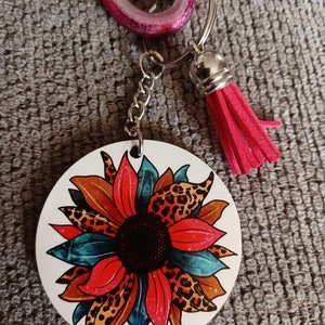 Op de afbeelding: Een ronde sleutelhanger met een kleurrijk zonnebloemontwerp. De zonnebloem heeft rode, blauwe en bruine bloemblaadjes met een zwart midden. De sleutelhanger is bevestigd aan een zilveren ketting en een roze kwast.