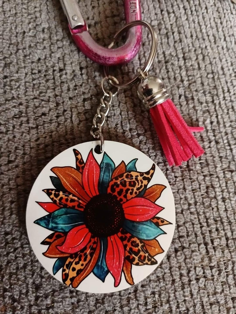 Op de afbeelding: Een ronde sleutelhanger met een kleurrijk zonnebloemontwerp. De zonnebloem heeft rode, blauwe en bruine bloemblaadjes met een zwart midden. De sleutelhanger is bevestigd aan een zilveren ketting en een roze kwast.