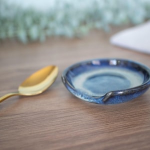 May include: A blue and white glazed ceramic spoon rest, alongside a gold-coloured spoon. The spoon rest has a curved design. The background is blurred, with a wooden surface.