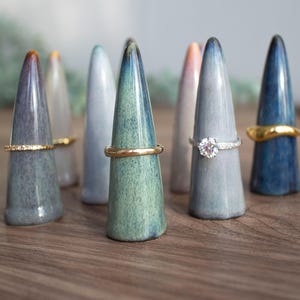 May include: A set of nine ceramic ring holders in various shades of blue and grey. Each holder is shaped like a cone and has a ring resting on top.