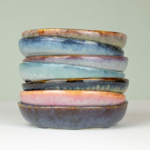 May include: A stack of eight small, round ceramic bowls in various shades of blue, brown, and pink. The bowls are glazed and have a slightly uneven surface.