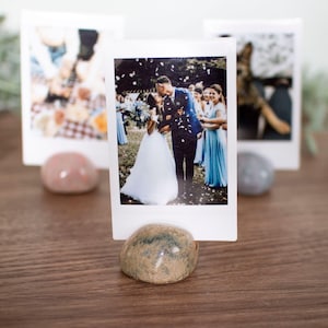 May include: A wedding photograph in a white frame, held by a small, speckled stone. Other photos are visible in the background, each held by a similar stone. The photos depict a wedding ceremony. The stones are on a wooden surface.