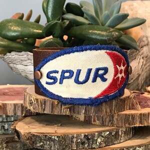 Op de afbeelding: Een bruin leren armband met een witte stoffen patch waarop "SPUR" in blauwe letters staat. De patch heeft een rood en blauw halvemaanontwerp met een witte ster.