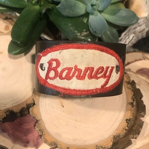 May include: A brown leather cuff bracelet with a white fabric patch that says "Barney" in red stitching.