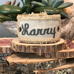 May include: A light brown fabric cuff bracelet with a frayed edge and a white canvas insert. The canvas insert has the name "Randy" embroidered in dark gray thread.