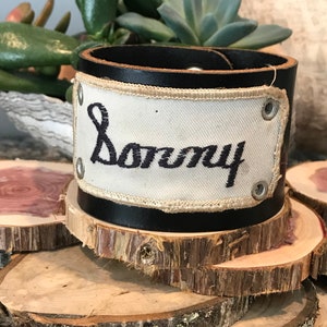 May include: A black leather cuff bracelet with a white canvas patch that says "Sonny" in black embroidery.
