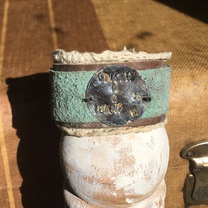 Könnte beinhalten: Dekoratives Holzobjekt mit weiß lackiertem Sockel und einem türkisgrünen Lederband. Das Band hat ein Metallschild mit der Aufschrift "Whiskey in a Teacup". Das Objekt ist mit einem Naturseil umwickelt.