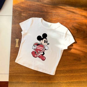 May include: White t-shirt featuring a cartoon Mickey Mouse holding a heart-shaped Valentine's Day greeting. The graphic includes red, black, and white colors. The text on the heart reads "Valentine Greetings".