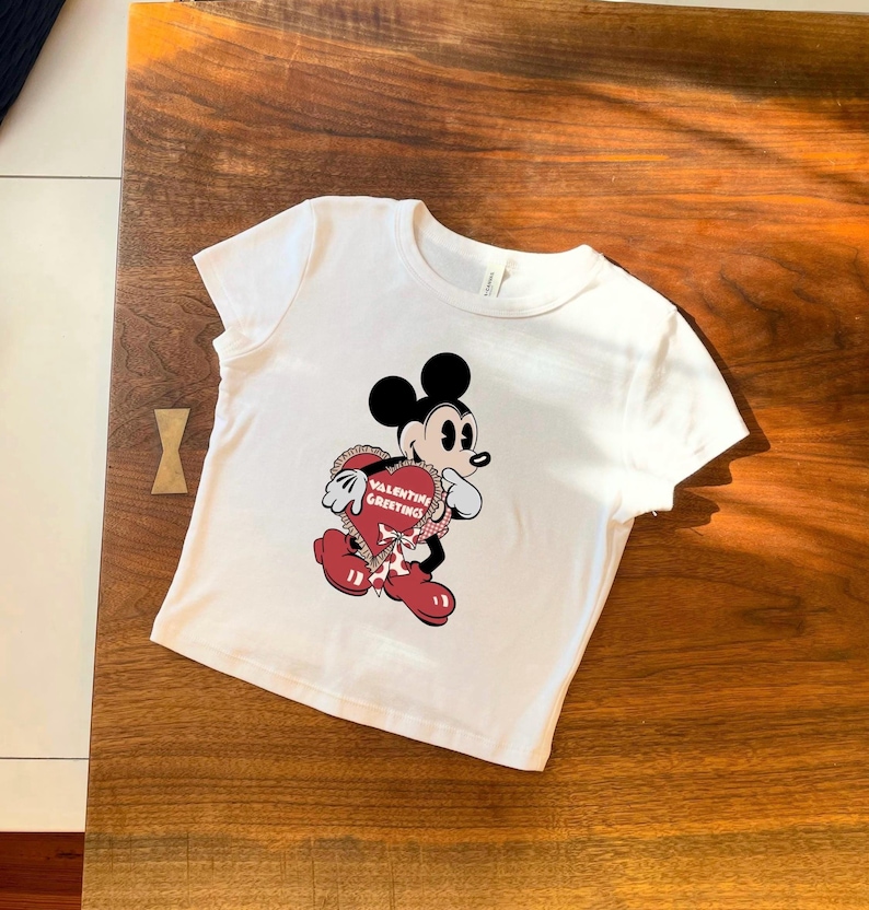 May include: White t-shirt featuring a cartoon Mickey Mouse holding a heart-shaped Valentine's Day greeting. The graphic includes red, black, and white colors. The text on the heart reads "Valentine Greetings".