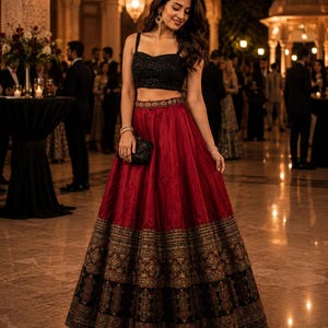 May include: A two-piece ensemble consisting of a black sleeveless crop top with a textured finish and a floor-length deep red skirt. The skirt features a wide, intricate gold and black patterned border at the hem and a matching decorative waistband.