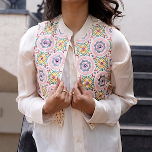 Può includere: Un gilet bianco ricamato con un motivo floreale colorato. Il gilet ha un colletto alla coreana e una chiusura con bottoni. Il gilet è indossato sopra una camicia bianca.