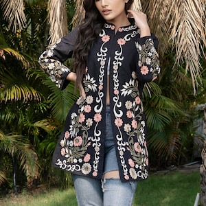 Peut inclure: Une veste noire brodée avec des motifs floraux en rose, blanc et vert. La veste a une longue ouverture à l'avant et est portée sur un jean bleu.